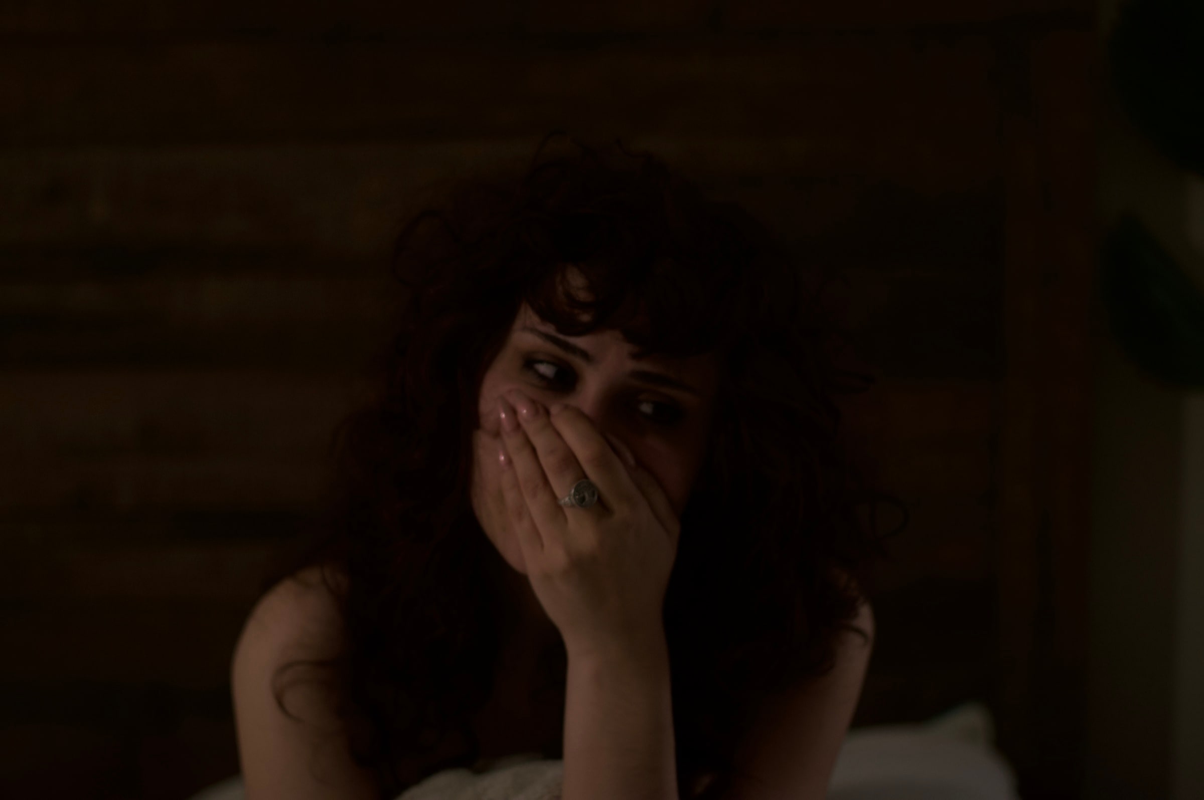 A curly-haired person holding their hands over their mouth in warm, low light.