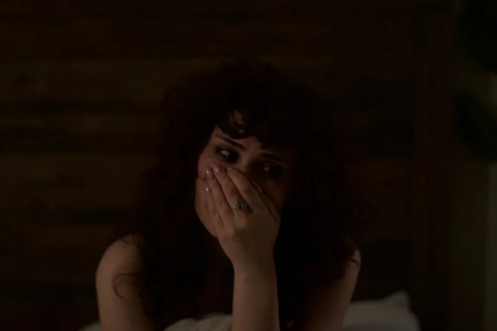 A curly-haired person holding their hands over their mouth in warm, low light.