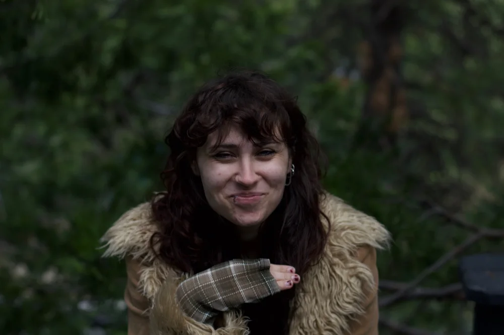 A person with curly hair smiling outdoors, wrapped in a scarf against green foliage.